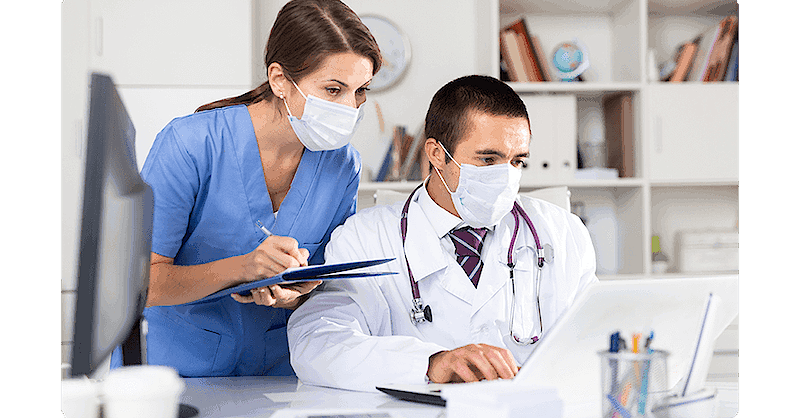 Two medical professionals with face masks sit looking at a laptop