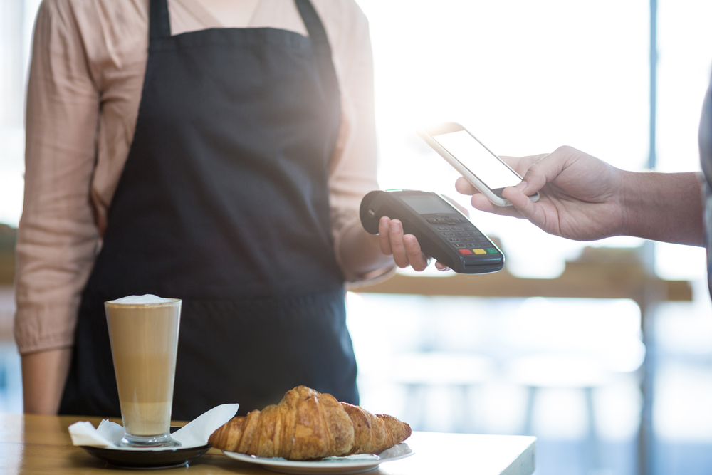 Barista holding payment terminal with croissant and coffee at a table